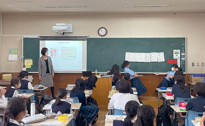 授業風景の写真。ふたりの生徒が黒板の前に出て、クラスのみんなに自分たちが並べた6枚のカードを見せている。柏野准教授とほかの生徒はカードに注目している