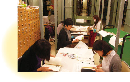 ４人の女性が図書館で書籍を調べている写真。机の上は資料でいっぱい。