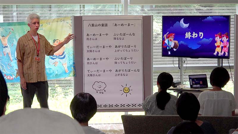 マシュー・トッピングさんがボードの横に立ち、童謡「あーめーまー」を解説している
