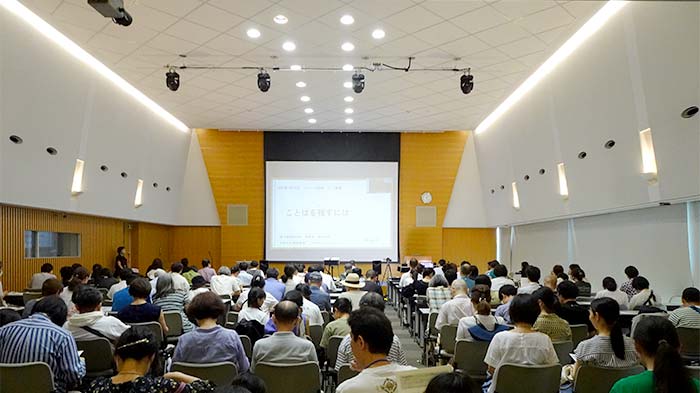 講堂に大勢の参加者が腰かけて講義が始まるのを待っている。