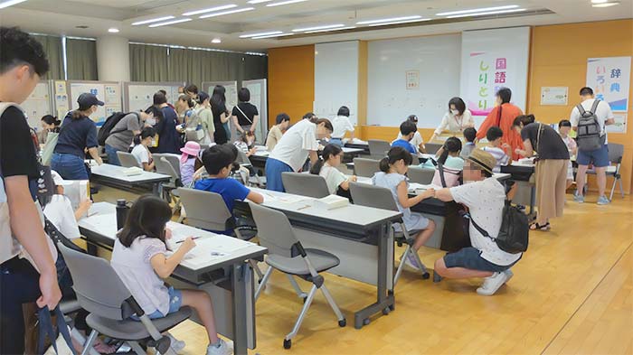 教室に机が並んでおり、多くの親子が辞書を開きながら問題を解いている写真